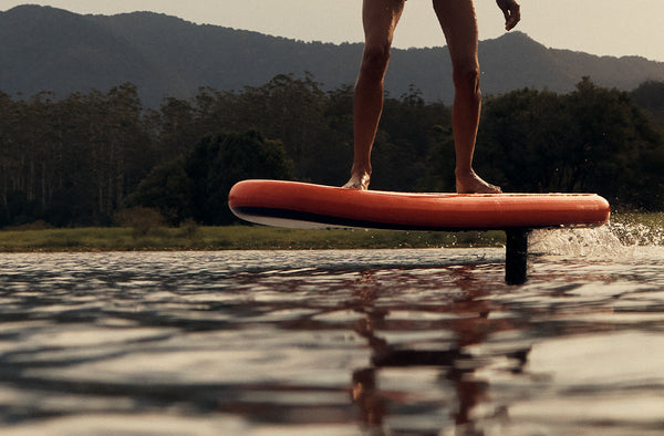 Close up view of a pair of legs standing on an eFoil board as it carves through a lake with mountains and trees in the background
